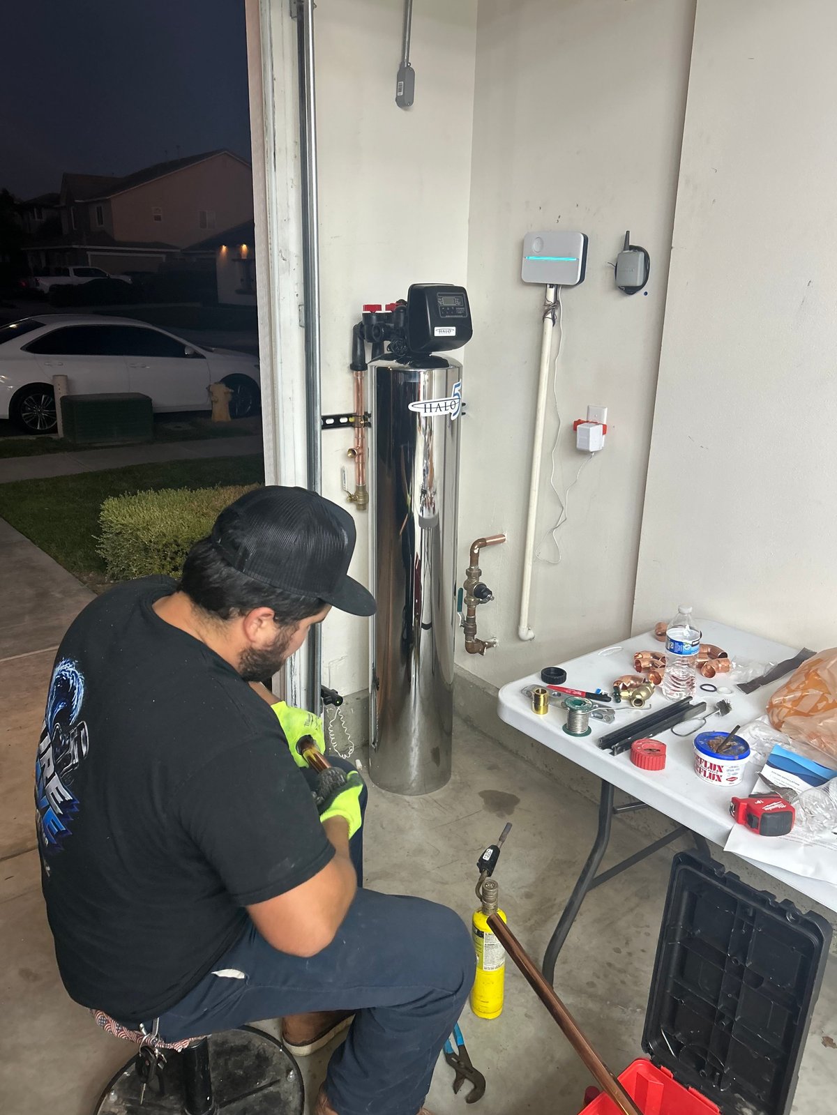 Technician in black shirt and cap servicing a water heater in a garage, with tools and equipment on a nearby table